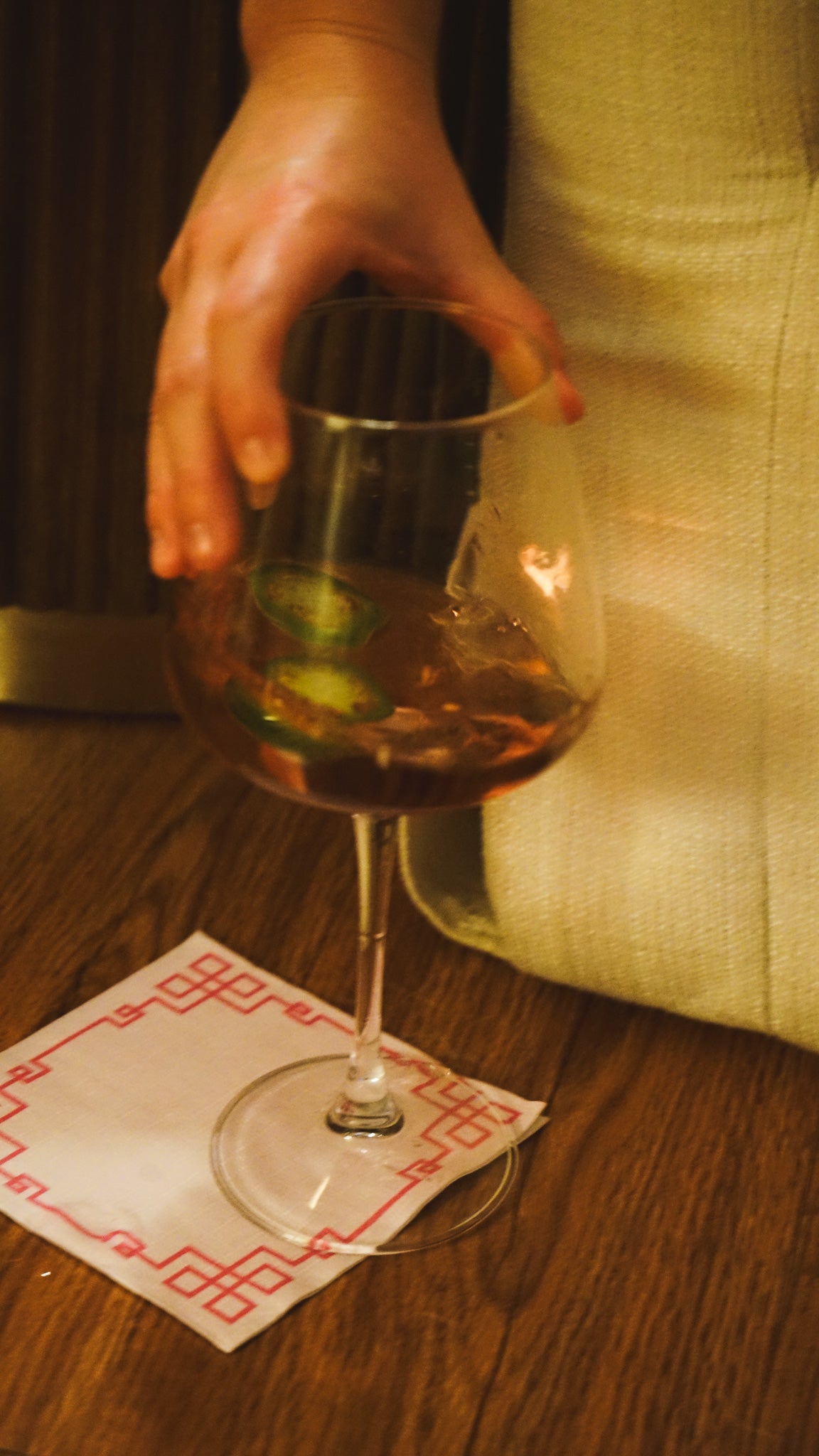 Hand holding a wine glass with a jalapeño slice on a wooden table.