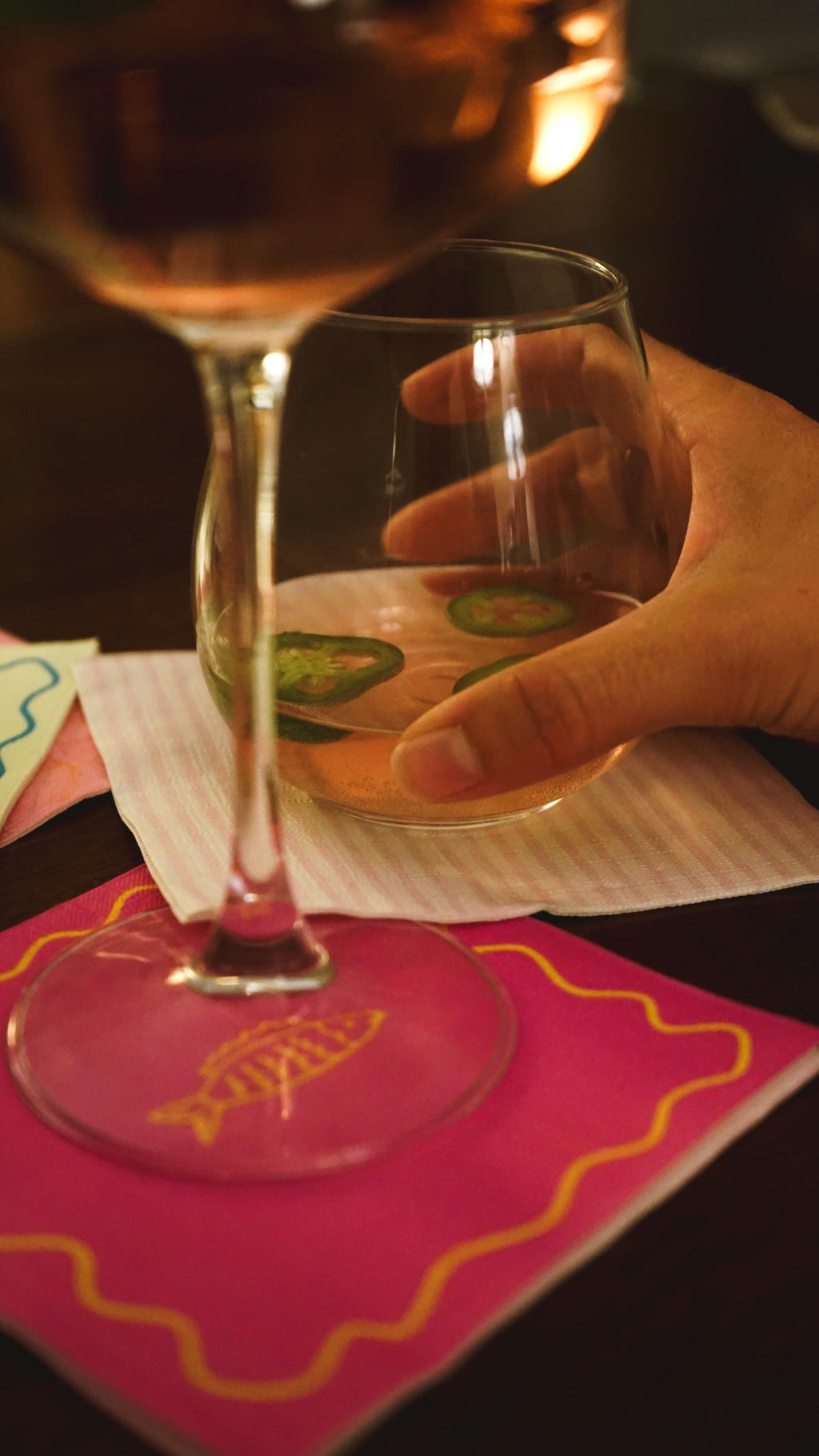 Hand holding a glass of white wine with a lime wedge on a pink coaster.