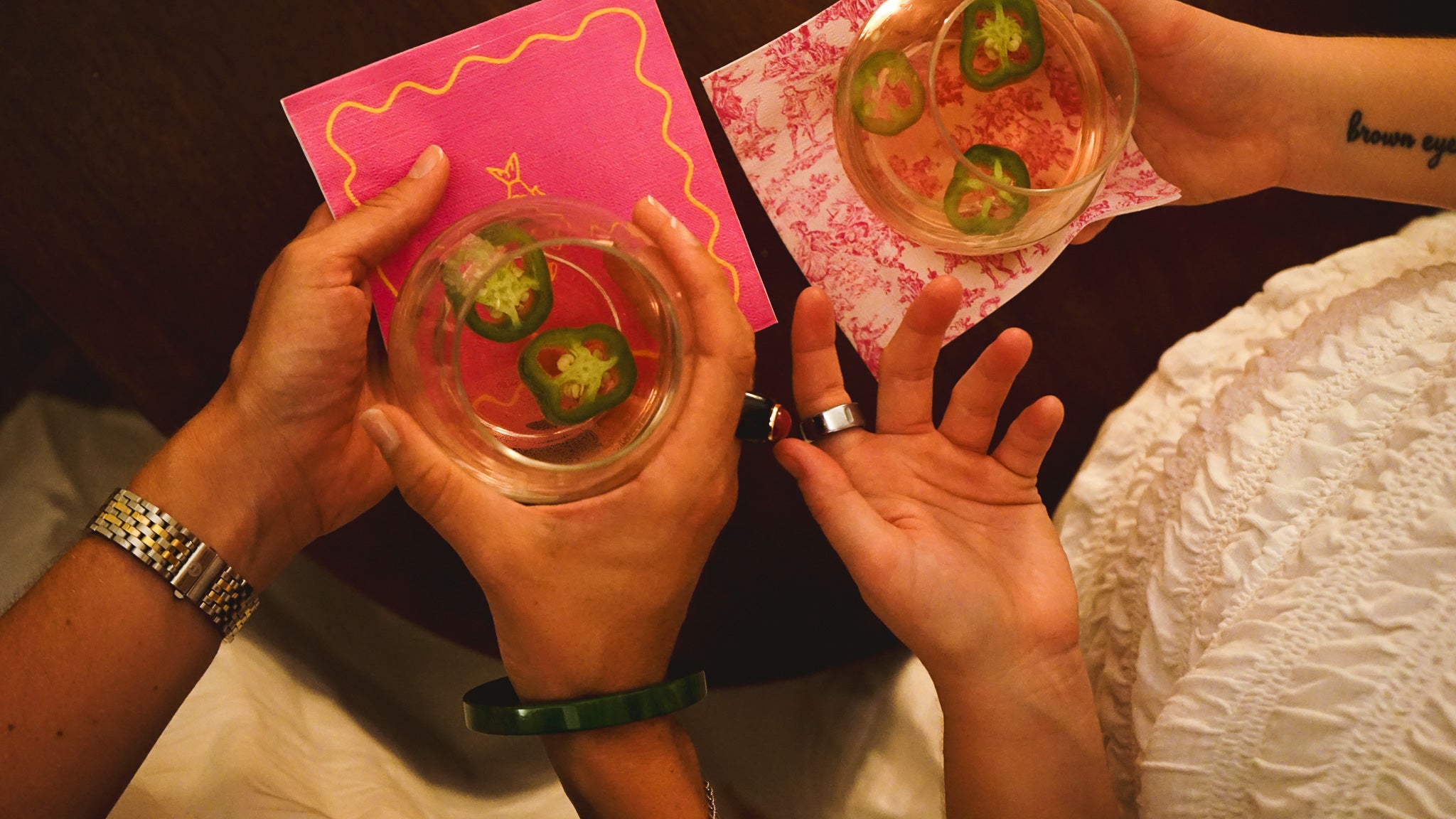 Two hands holding small cups with green slices, one on a pink card, against a dark background.